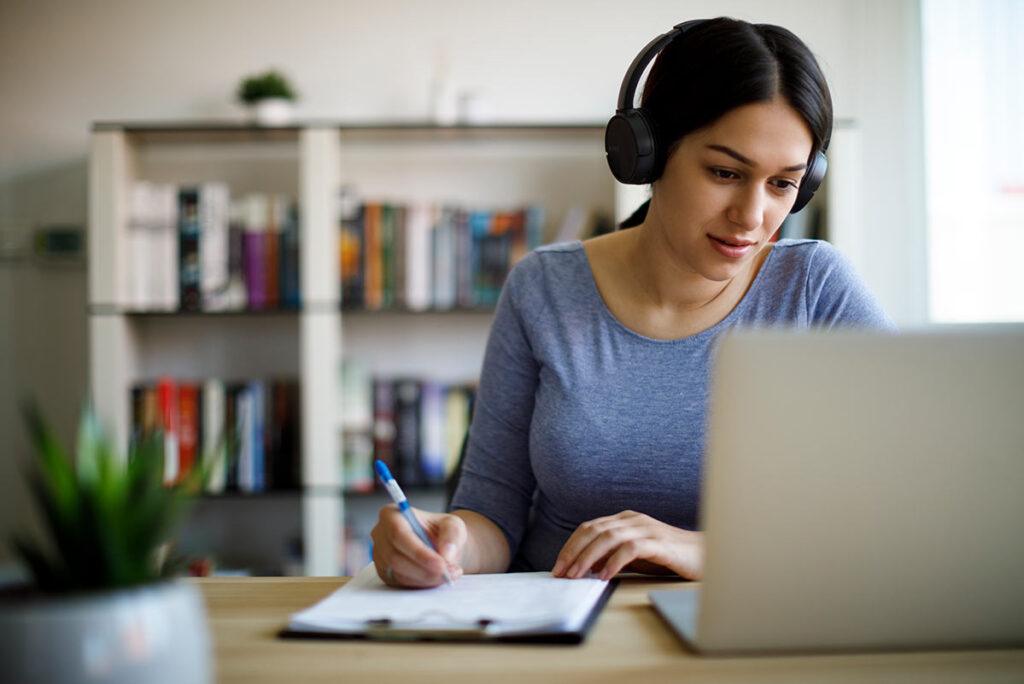 Woman with headphones on sitting at laptop and writing on clipboard